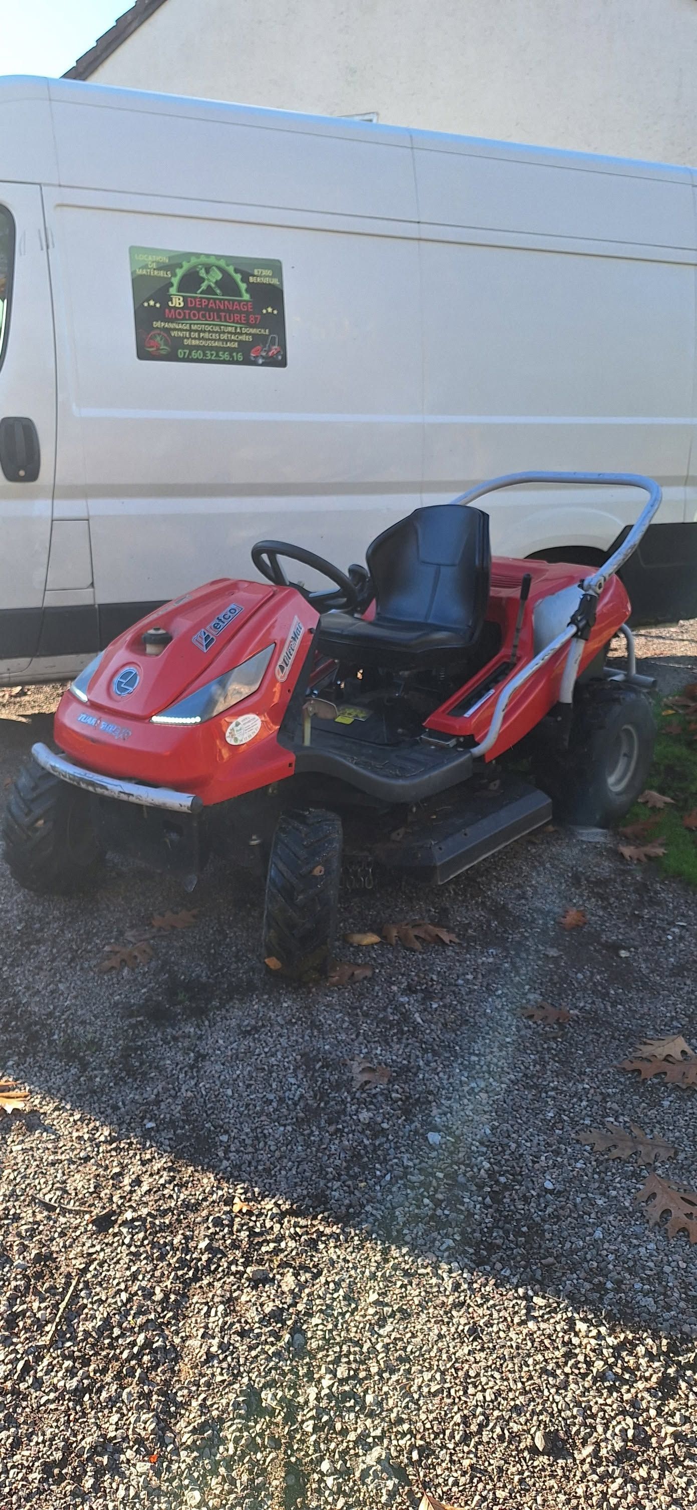 Tondeuse à gazon autoportée rouge garée sur du gravier à côté d'une camionnette blanche.