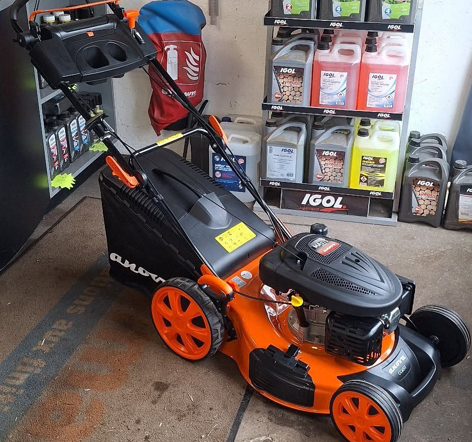 Tondeuse à gazon BLACK+DECKER orange et noire dans un garage, à côté d'étagères remplies d'huile et de fluides.