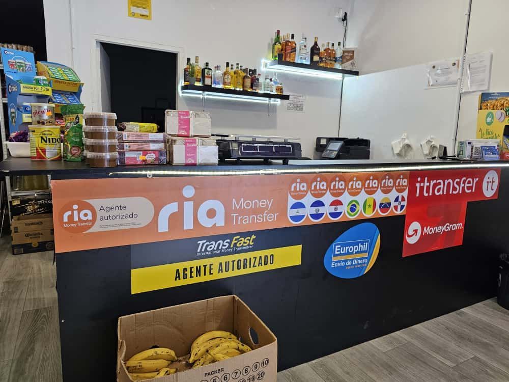Un mostrador de caja registradora con publicidad de la empresa de transferencia de dinero Ria y una caja de plátanos. Estanterías de licores al fondo.