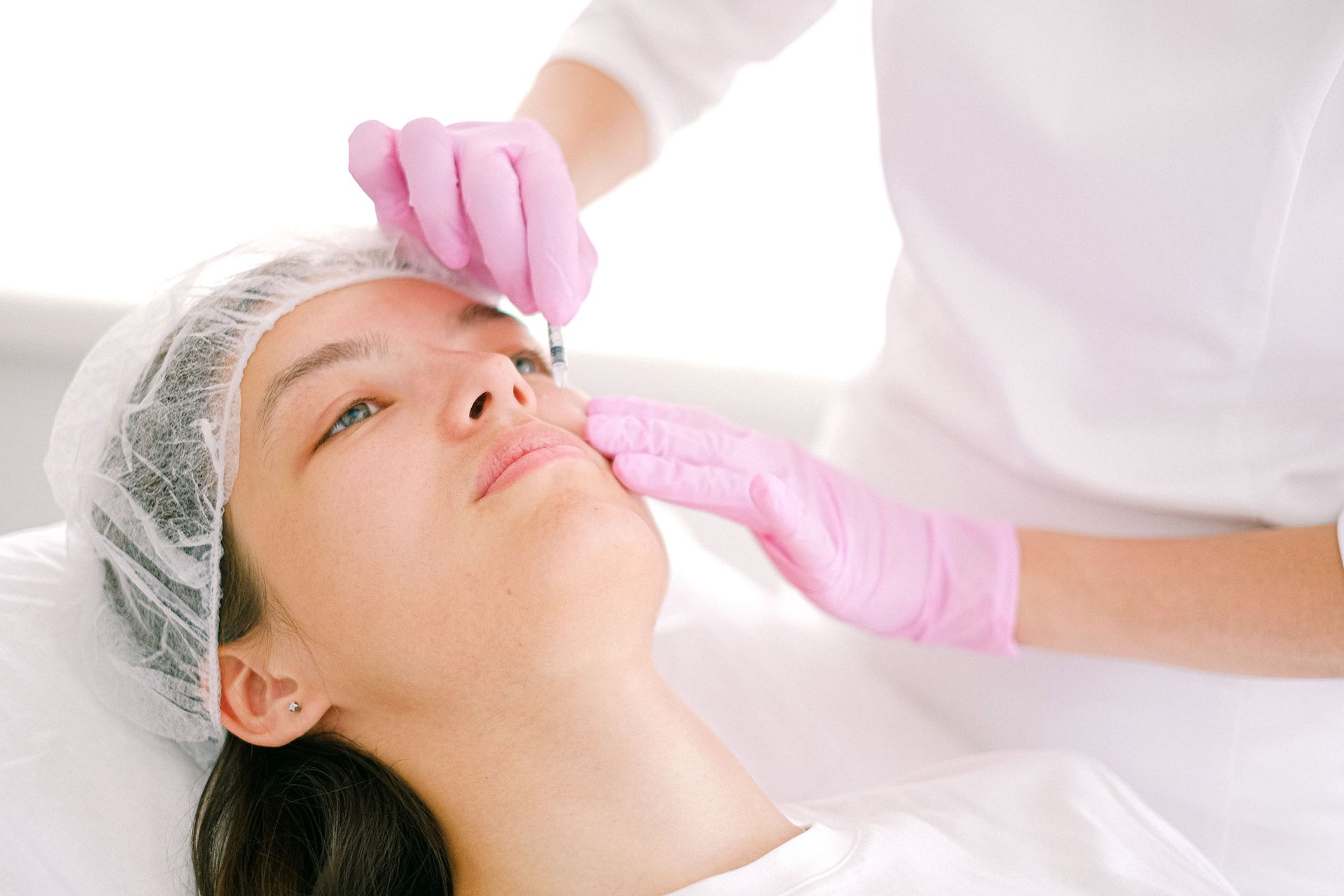 Tratamiento cosmético en el rostro de una mujer con manos enguantadas de rosa en una clínica luminosa.