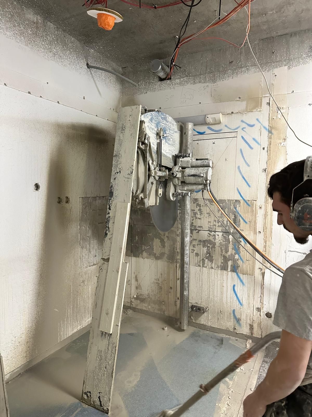 Un homme utilise une scie murale dans une pièce poussiéreuse, coupant du béton, portant une protection auditive.