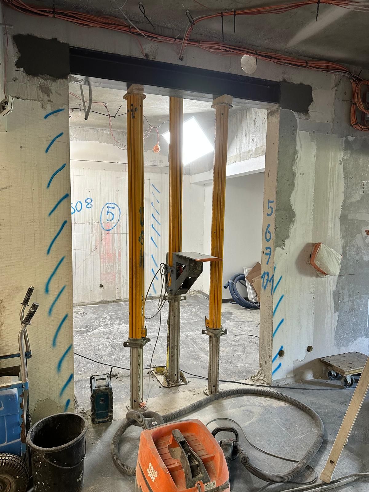 Chantier de construction avec poutres de soutènement dans une porte. Des vérins jaunes soutiennent le plafond pendant les travaux.