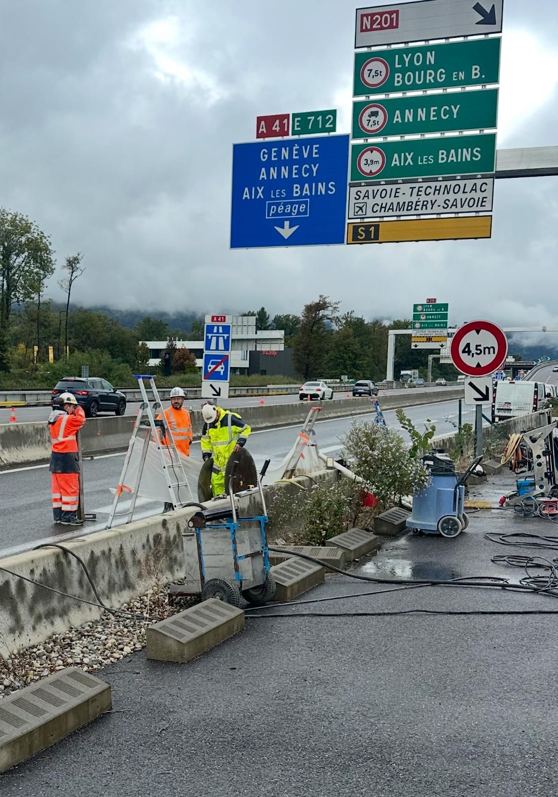 Des ouvriers réparent une barrière sur une autoroute, avec des panneaux indiquant Lyon, Annecy et Aix-les-Bains. Jour couvert.