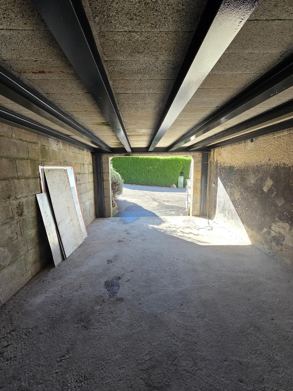 Abri voiture intérieur en béton, ouvert sur une rue ensoleillée. Des poutres noires traversent le plafond.