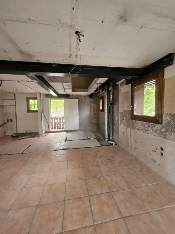 Pièce intérieure en cours de rénovation ; poutres apparentes, carrelage au sol, fenêtres, murs bruts.