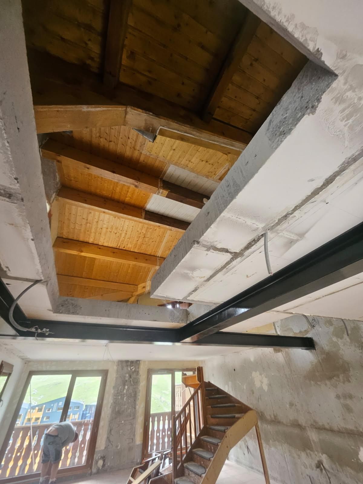 Vue intérieure d'un bâtiment en construction, présentant des poutres en bois apparentes, du béton et un escalier.