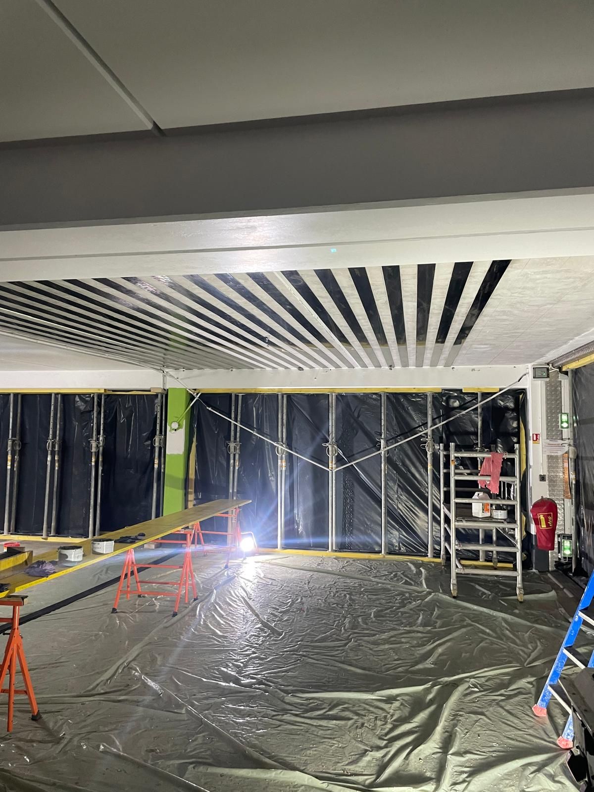 Construction en cours : une pièce avec un plafond à rayures noires, une bâche en plastique au sol et une échelle.
