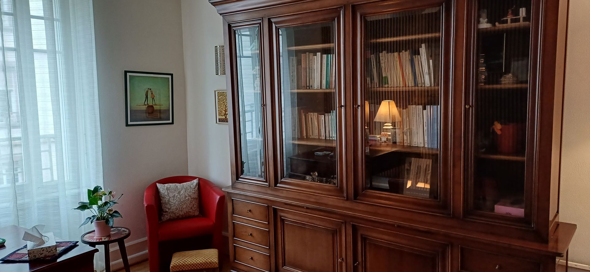 Une armoire en bois aux portes vitrées remplie de livres, une chaise et une fenêtre avec des rideaux.