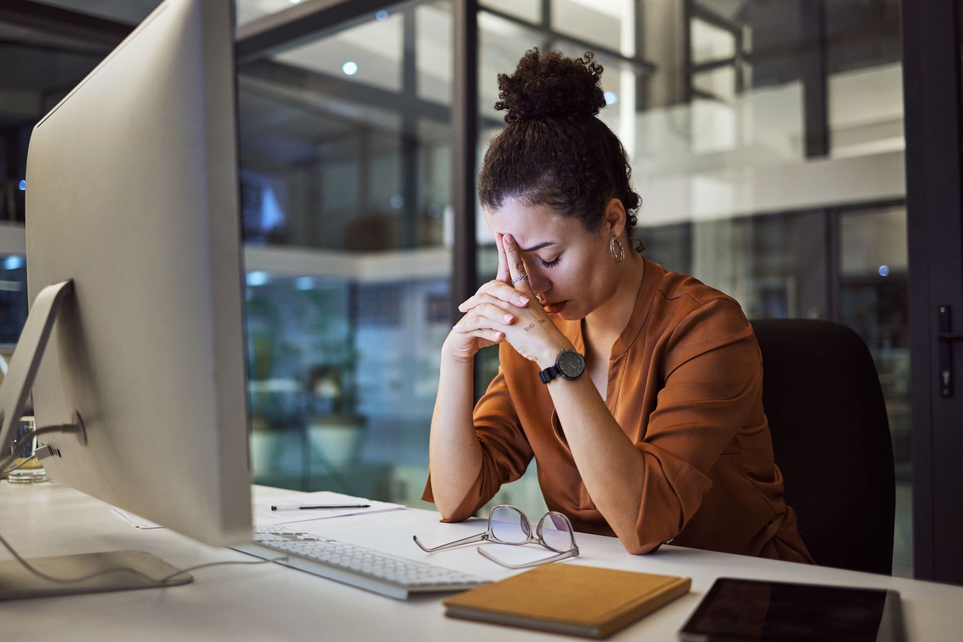 Femme aux cheveux bouclés, les mains sur le visage, l'air stressé devant un ordinateur dans un bureau, le soir.