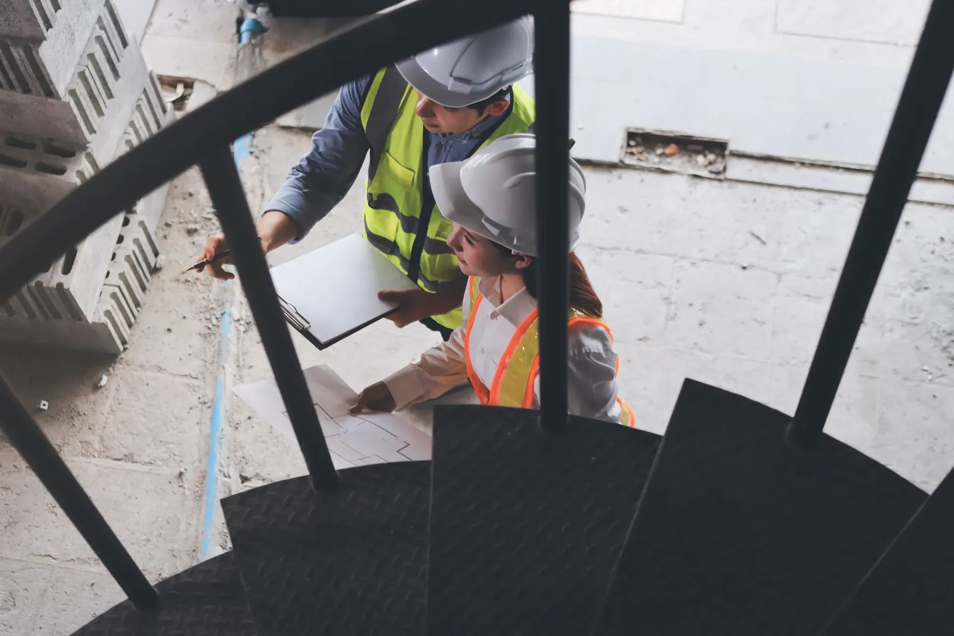 Dos personas con cascos y chalecos en una escalera de caracol, revisando planos en un sitio de construcción.