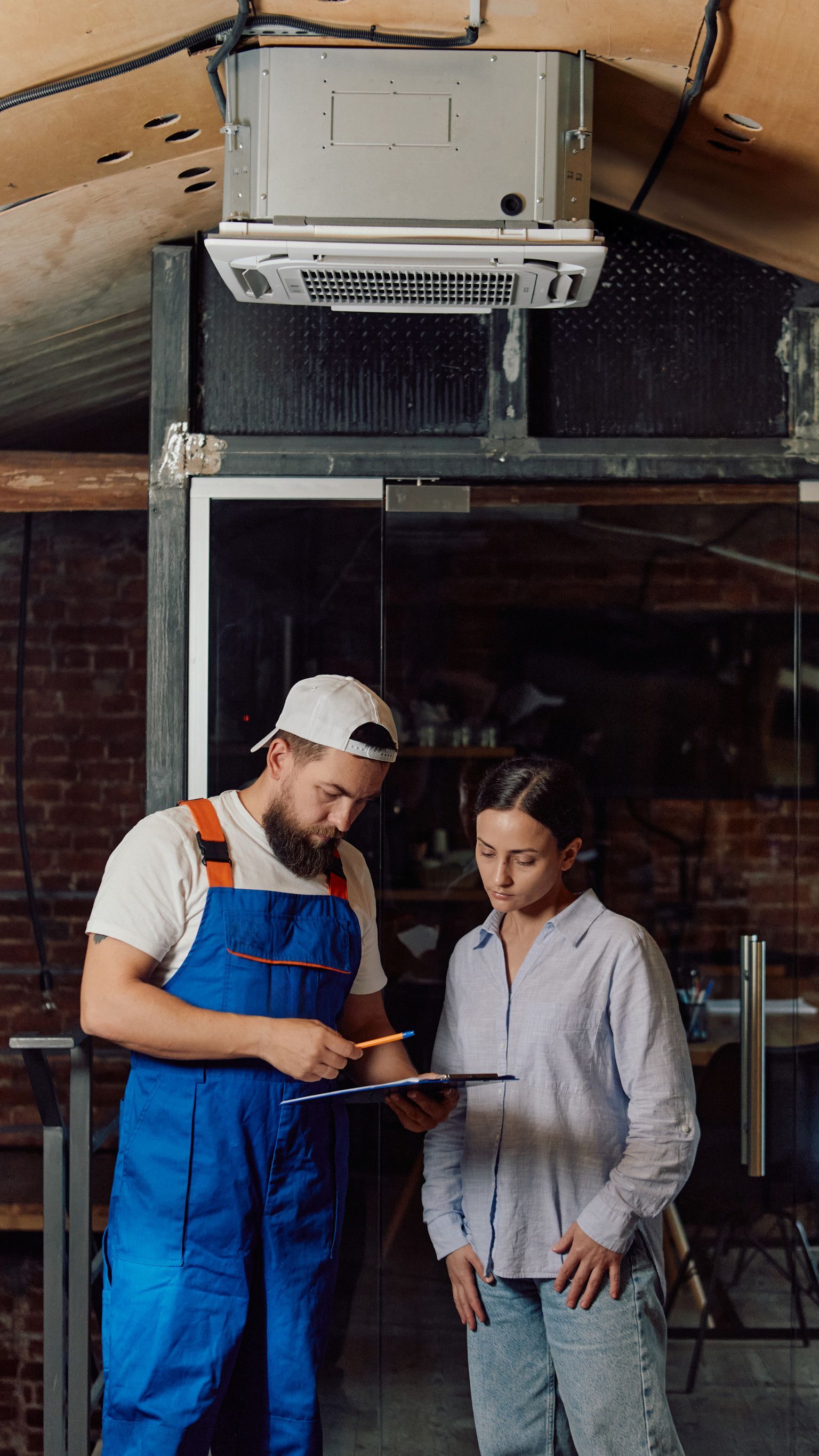 Mecánico con mono azul y una mujer mirando un portapapeles cerca de una unidad de aire acondicionado.