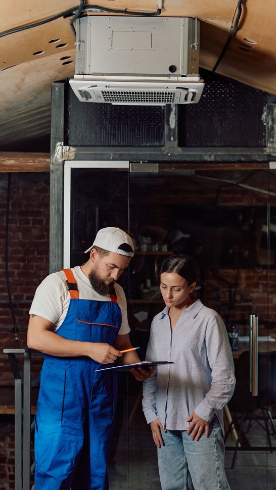 Mecánico con mono azul y una mujer mirando un portapapeles cerca de una unidad de aire acondicionado.