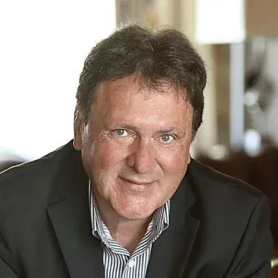 Nahaufnahme eines lächelnden Mannes in einem dunklen Anzugjackett und einem gestreiften Hemd, aufgenommen in einem Innenraum.