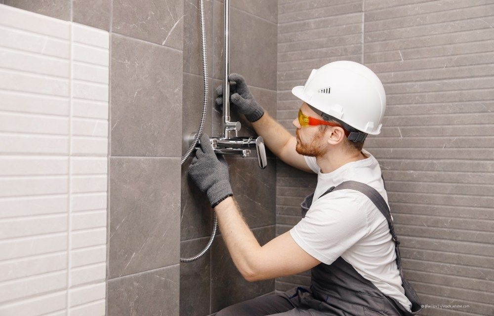 Ein Arbeiter montiert eine Duscharmatur an einer grau gefliesten Badezimmerwand