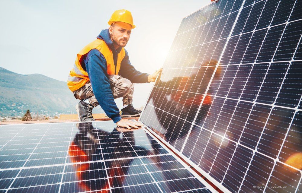 Arbeiter in orangefarbener Schutzkleidung installiert Solarpaneele auf einem Dach im hellen Sonnenlicht