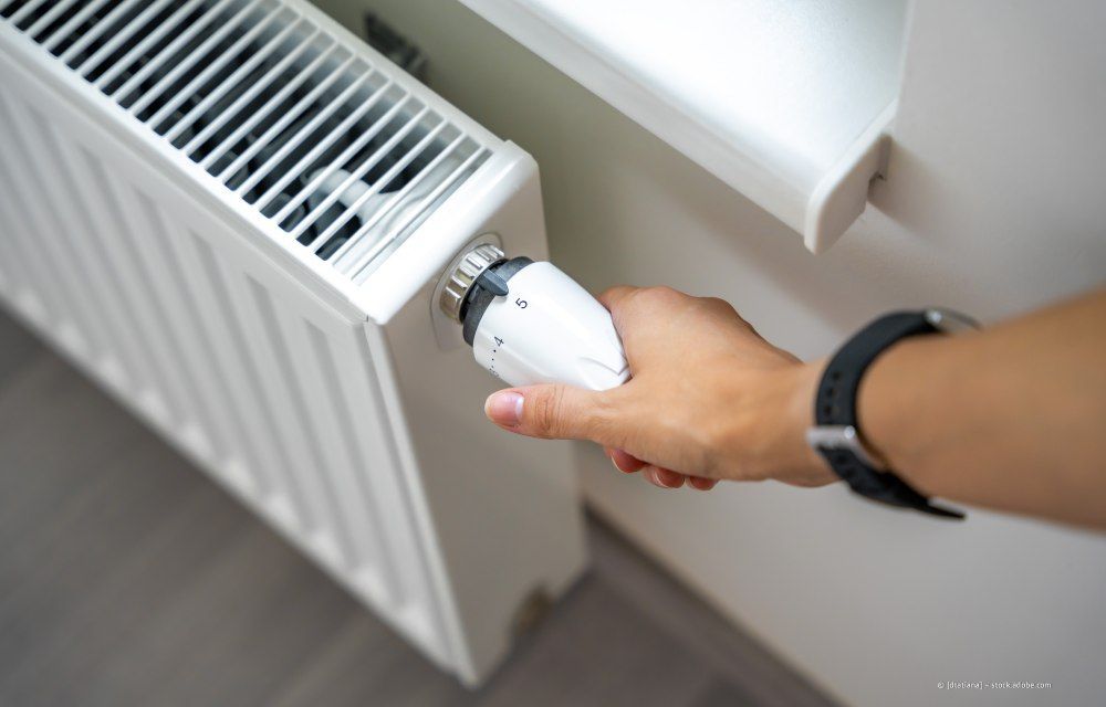 Hand dreht ein weißes Thermostatventil an einem Heizkörper in der Nähe eines Fensters
