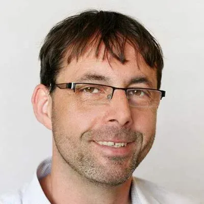 Porträt einer lächelnden Person mit Brille und weißem Hemd vor einfarbigem Hintergrund