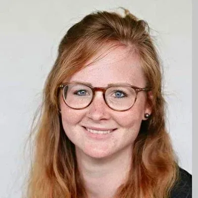 Lächelnde Person mit Brille und roten Haaren vor hellem Hintergrund