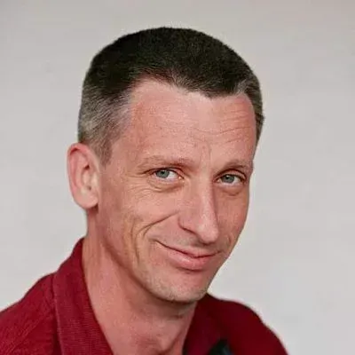 Nahaufnahme einer Person mit kurzen dunklen Haaren, einem leichten Lächeln und einem roten Hemd vor einem einfarbigen hellen Hintergrund