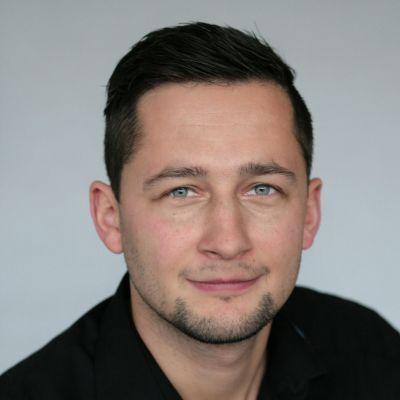 Studioaufnahme eines Mannes mit kurzen dunklen Haaren und hellen Augen, der ein schwarzes Hemd trägt, vor grauem Hintergrund