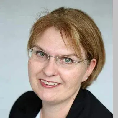 Lächelnde Person mit kurzen hellbraunen Haaren und Brille, die ein schwarzes Oberteil trägt, vor hellgrauem Hintergrund