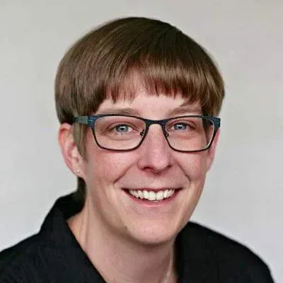 Person mit kurzen braunen Haaren und Brille lächelt vor hellem Hintergrund