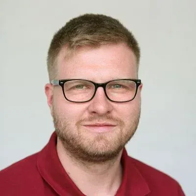 Porträt eines Mannes mit schwarzer Brille und rotem Poloshirt vor hellgrauem Hintergrund