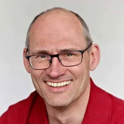 Lächelnder, glatzköpfiger Mann mit Brille und rotem Hemd vor hellem Hintergrund
