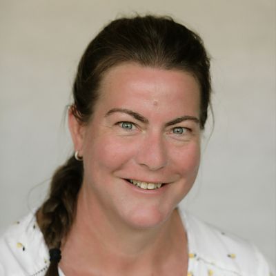 Lächelnde Person mit dunklen Haaren in einer weißen Bluse vor einem einfarbigen hellen Hintergrund