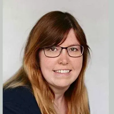 Porträt einer lächelnden Person mit Brille und braunem Haar vor einem einfarbigen, hellen Hintergrund