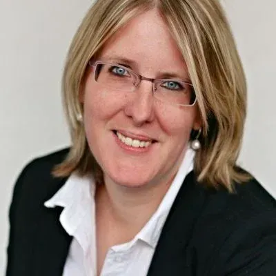 Porträt einer lächelnden Person mit Brille, die einen schwarzen Blazer und ein weißes Hemd trägt.