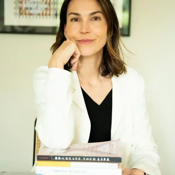 Mujer con blazer blanco, con la barbilla apoyada en la mano, sonriendo. Libros apilados delante.