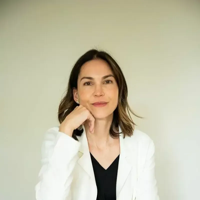 Mujer con blazer blanco, con la barbilla apoyada en la mano, sonriendo. Libros apilados delante.