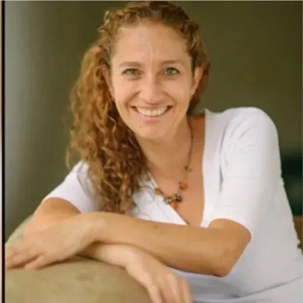 Mujer de cabello rizado sonríe, con los brazos cruzados, apoyada sobre una superficie de color claro.