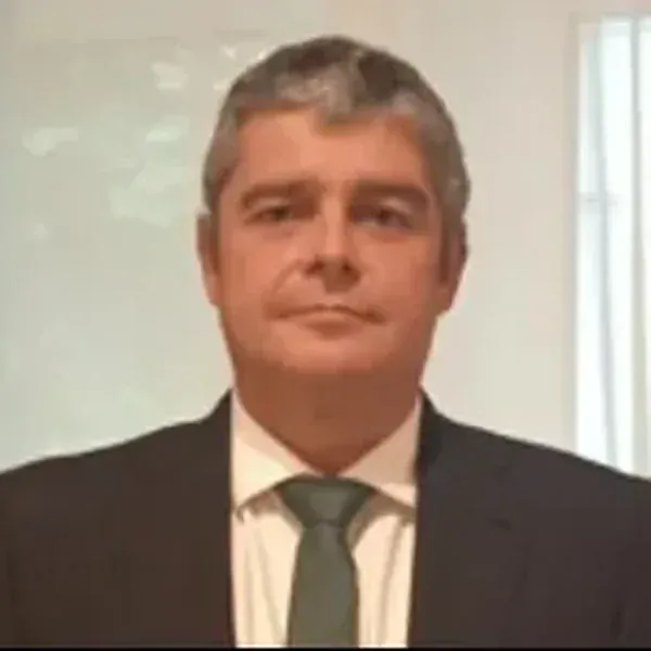 Hombre con traje oscuro, camisa blanca, corbata verde, mirando hacia adelante, expresión neutra.