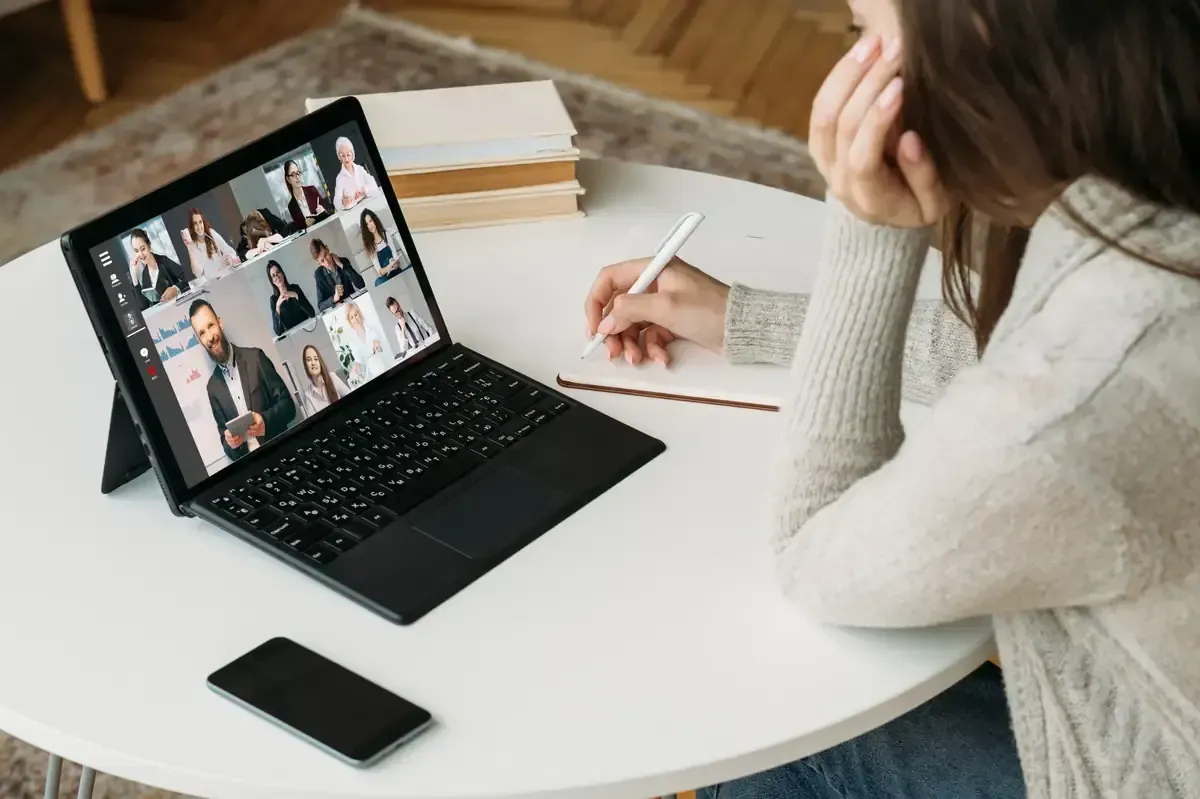 Mujer en una mesa en una videoconferencia en una computadora portátil, tomando notas.