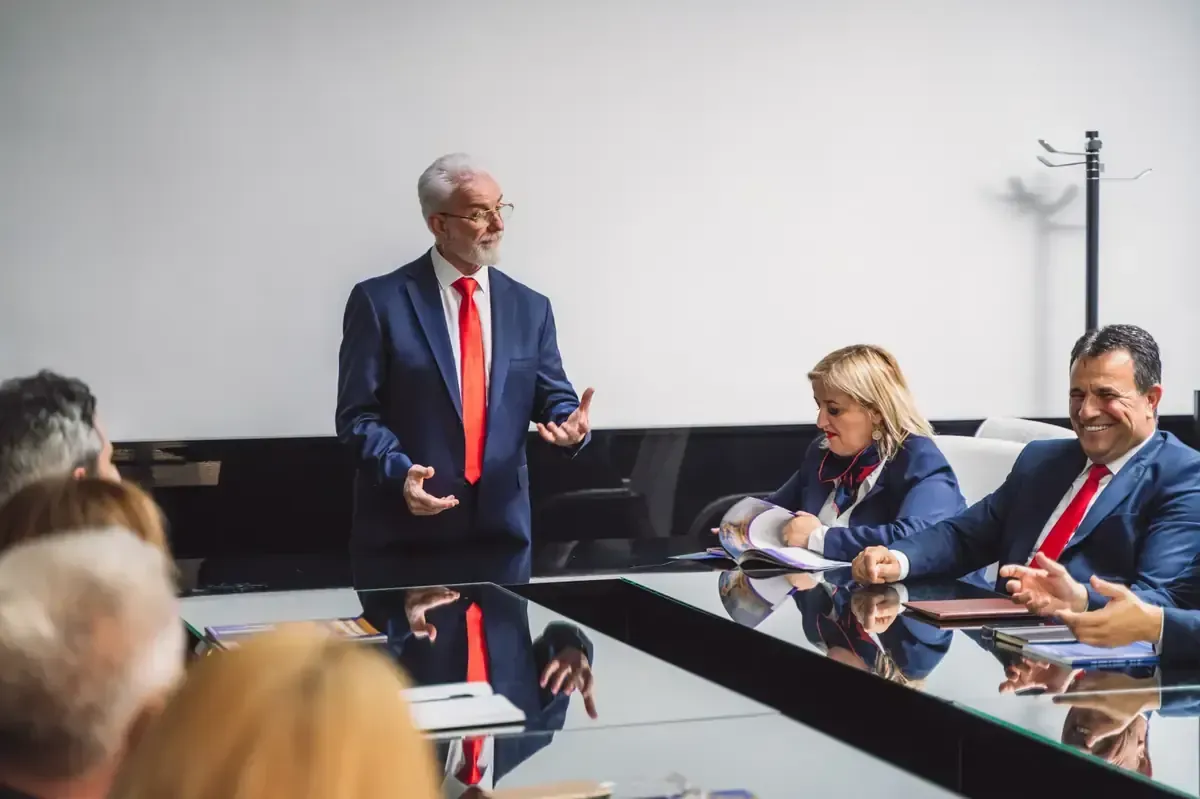 Hombre de traje dando una presentación en una mesa de conferencias, mientras otros escuchan y toman notas.