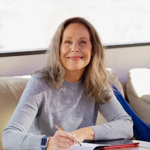 Mujer de cabello largo y canoso escribe en una mesa, sonriendo. Lleva un suéter gris y un reloj.