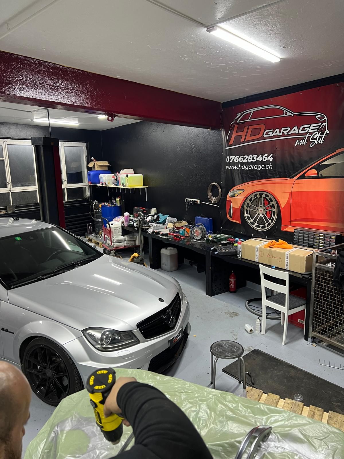 Silberner Mercedes in einer Garage, eine Person hält eine Bohrmaschine, Werkzeuge und Autoteile sind zu sehen.