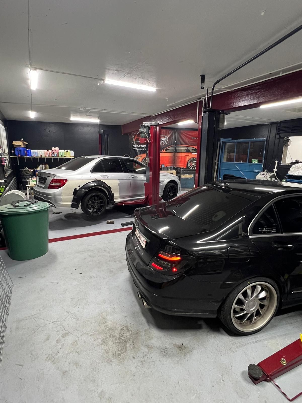 Eine Autowerkstatt mit zwei Autos. Eines schwarz, eines weiß, mit ausgebauten Teilen. Rote Hebebühne und Werkzeuge.