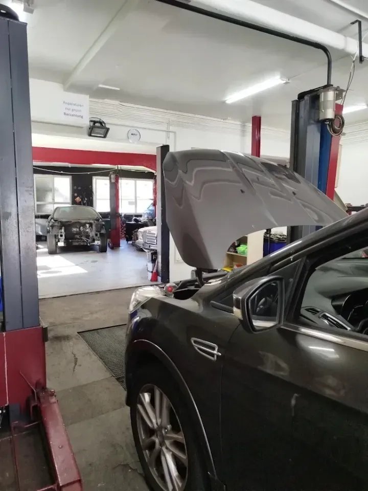 Auto in Autowerkstatt mit geöffneter Motorhaube, Hebebühnen auf beiden Seiten. Andere Autos in der Werkstatt sichtbar, weiße und rote Wände.