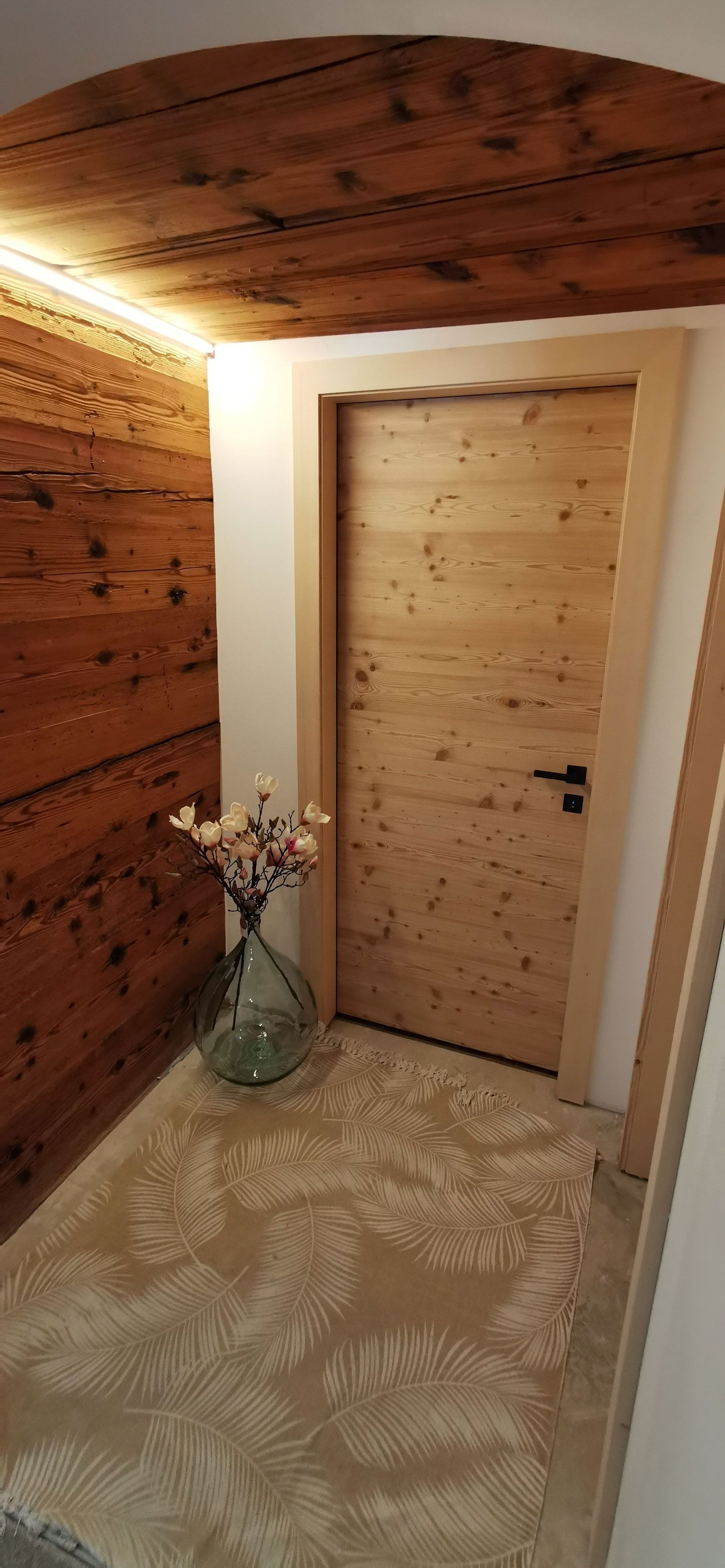 Un couloir avec une porte en bois et un vase de fleurs au sol.