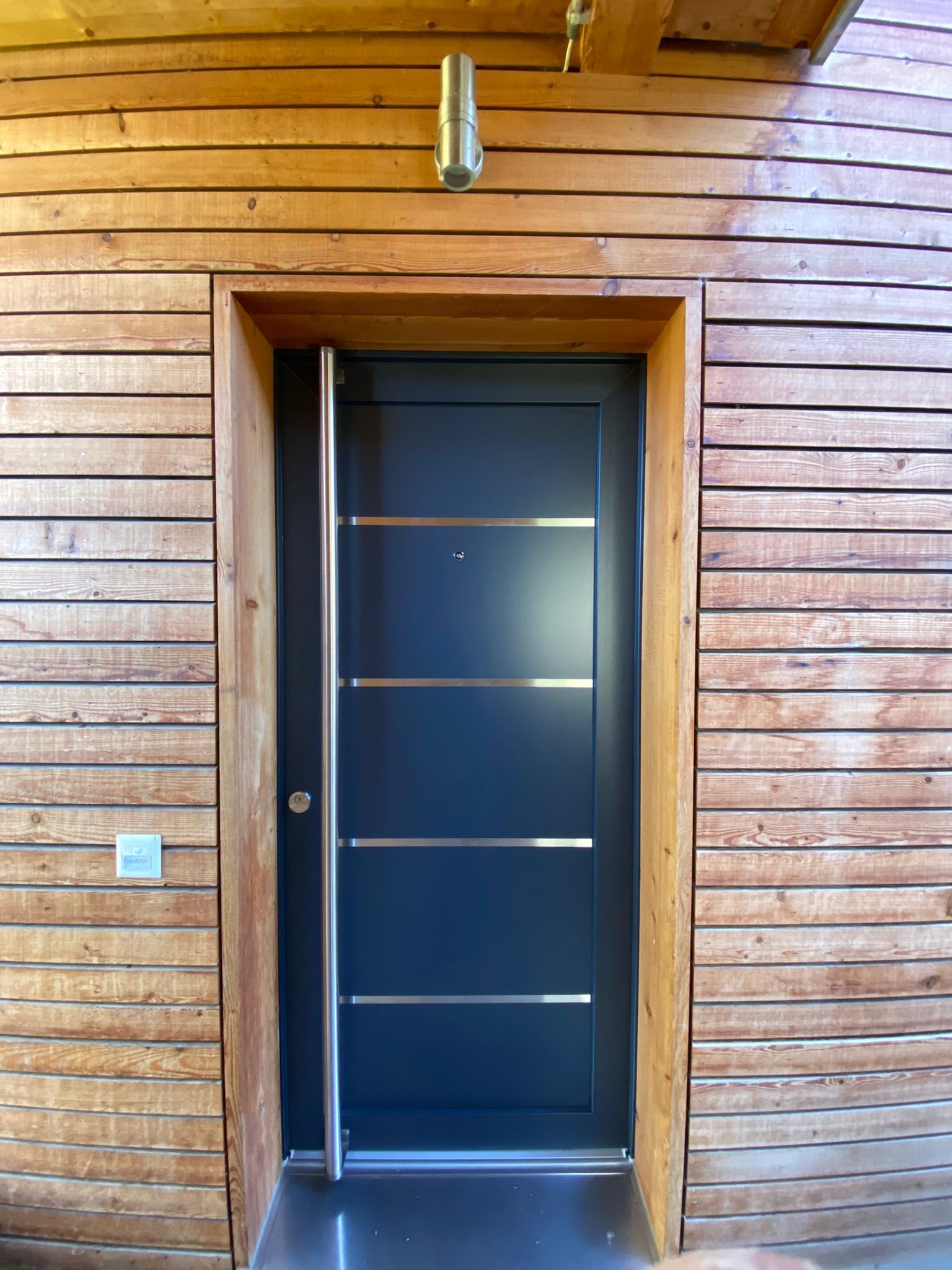 Une porte noire avec garniture en acier inoxydable se trouve sur un bâtiment en bois.