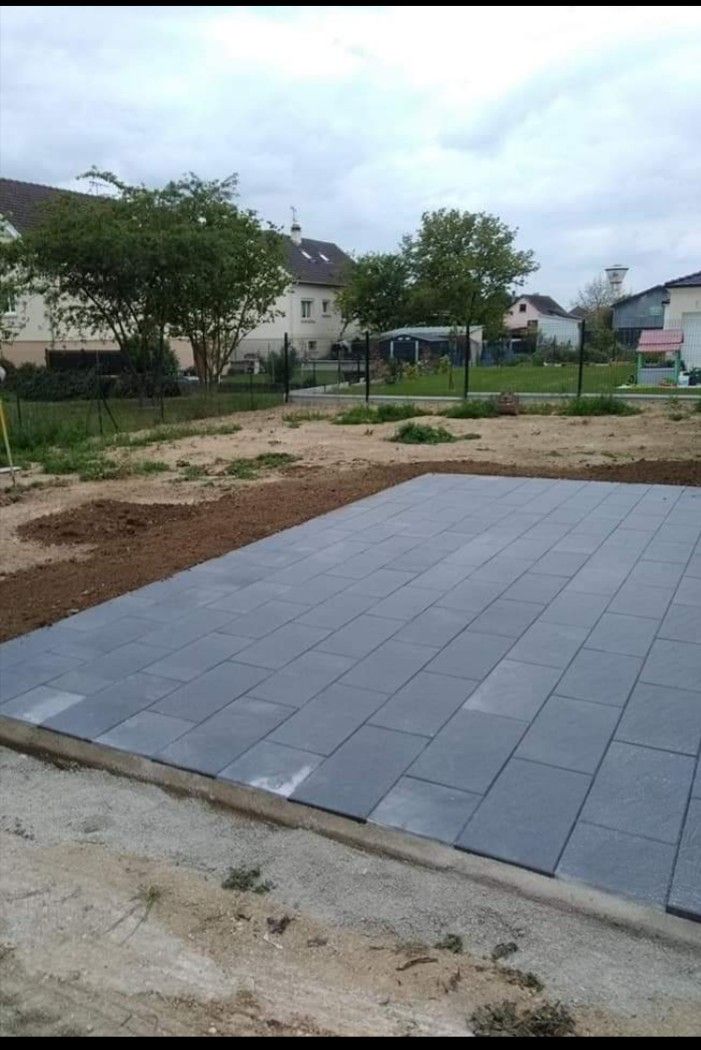 Terrasse en dalles grises pavées en construction dans un jardin sablonneux avec des maisons et des arbres en arrière-plan