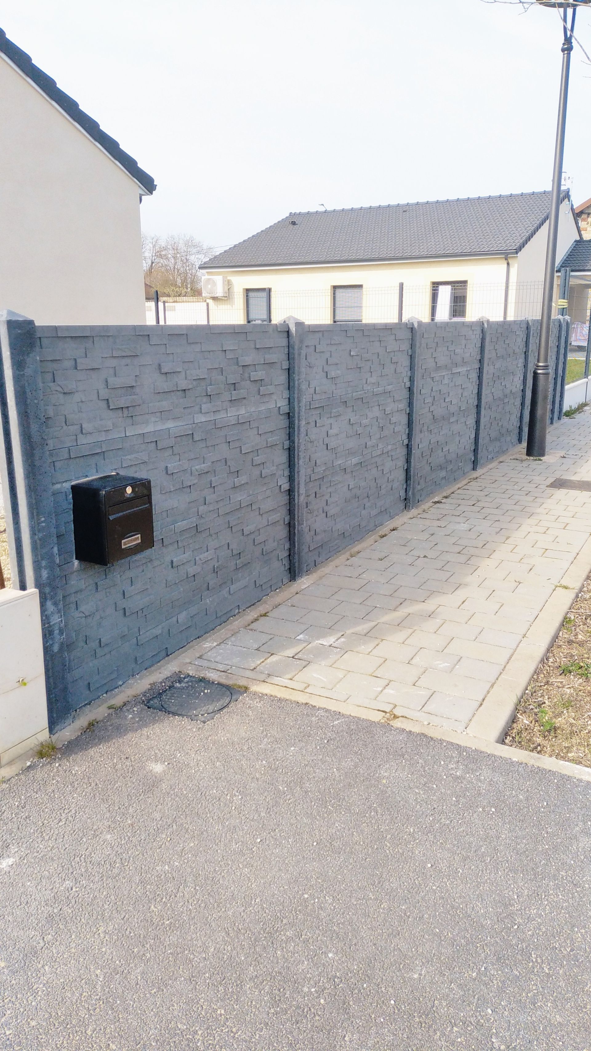 Mur en béton gris longeant un trottoir en pente à côté d'une maison, avec une boîte aux lettres et une allée pavée.