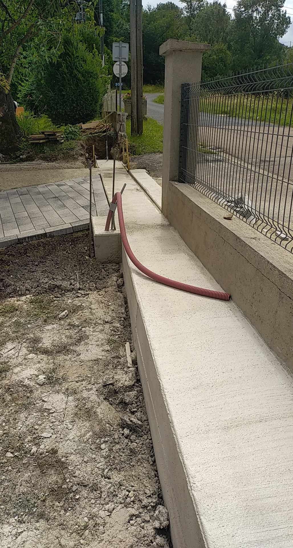 Allée en ciment longeant un mur en béton, avec un tuyau ou un câble rouge couché sur le bord près d'un portail.