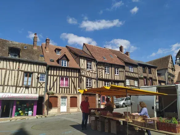 Un marché ensoleillé en plein air, avec des bâtiments historiques à ossature bois en arrière-plan et un étal jaune au premier plan.
