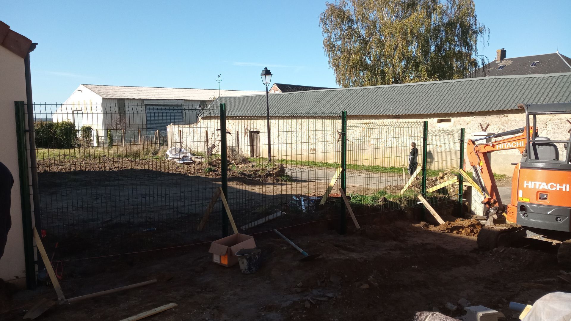 Une excavatrice travaille sur un chantier de construction où plusieurs poteaux de clôture métalliques verticaux sont installés dans le sol.