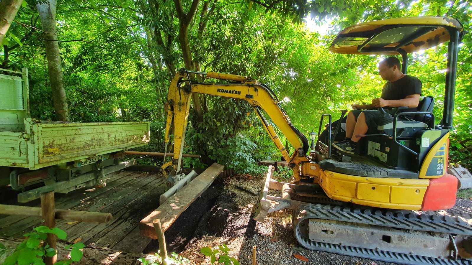 Une personne manœuvre une minipelle jaune pour déplacer des poutres en bois près d'une remorque dans un environnement boisé en extérieur.