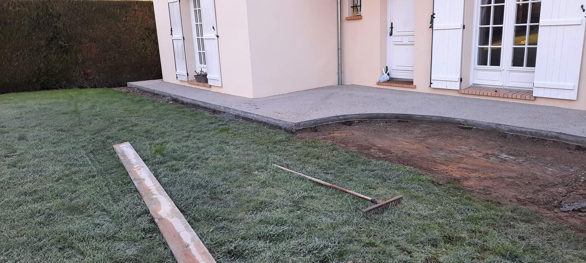 L'extérieur d'une maison avec une terrasse pavée et une partie en terre ratissée dans une cour gazonnée, avec un râteau posé sur la pelouse.
